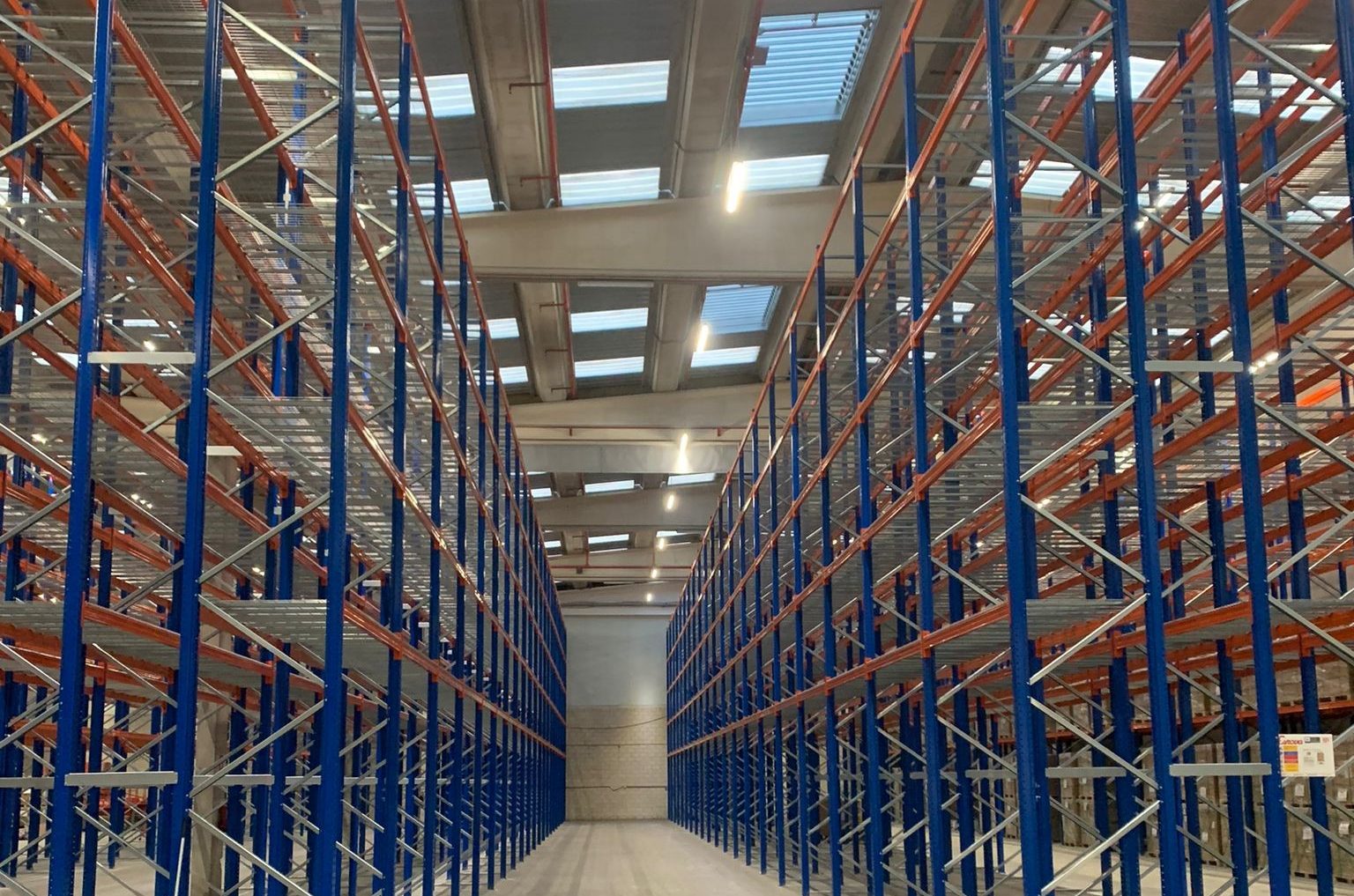 Empty warehouse with tall blue and orange metal storage racks under a ceiling with skylights and fluorescent lights.