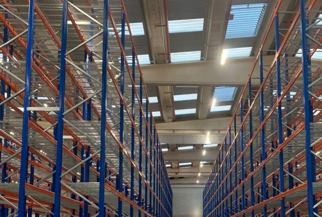 Empty warehouse with tall blue and orange metal storage racks under a ceiling with skylights and fluorescent lights.