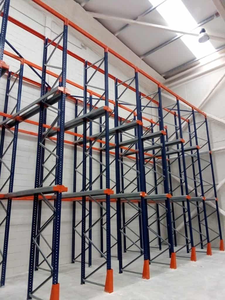 Empty industrial storage racks with blue frames and orange supports are installed against a white brick wall in a warehouse with a high ceiling.