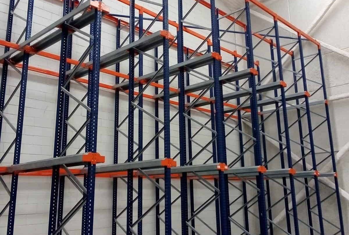 Empty industrial storage racks with blue frames and orange supports are installed against a white brick wall in a warehouse with a high ceiling.
