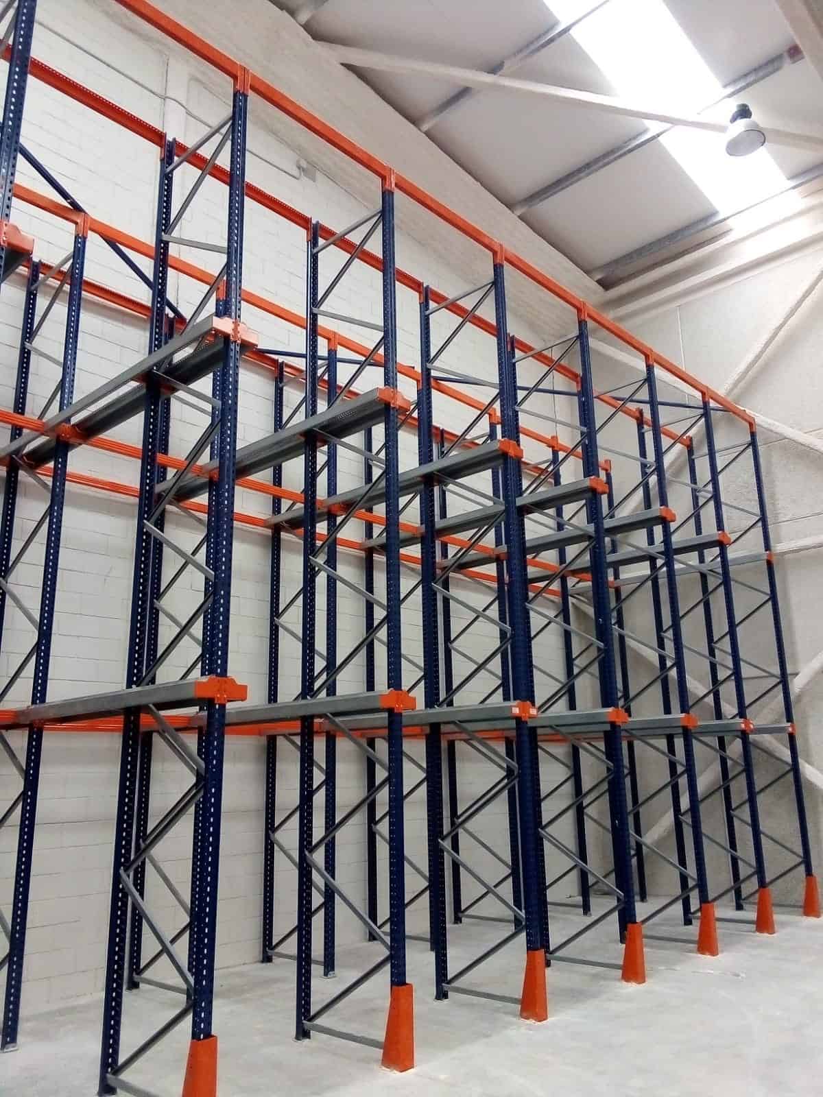 Empty industrial storage racks with blue frames and orange supports are installed against a white brick wall in a warehouse with a high ceiling.