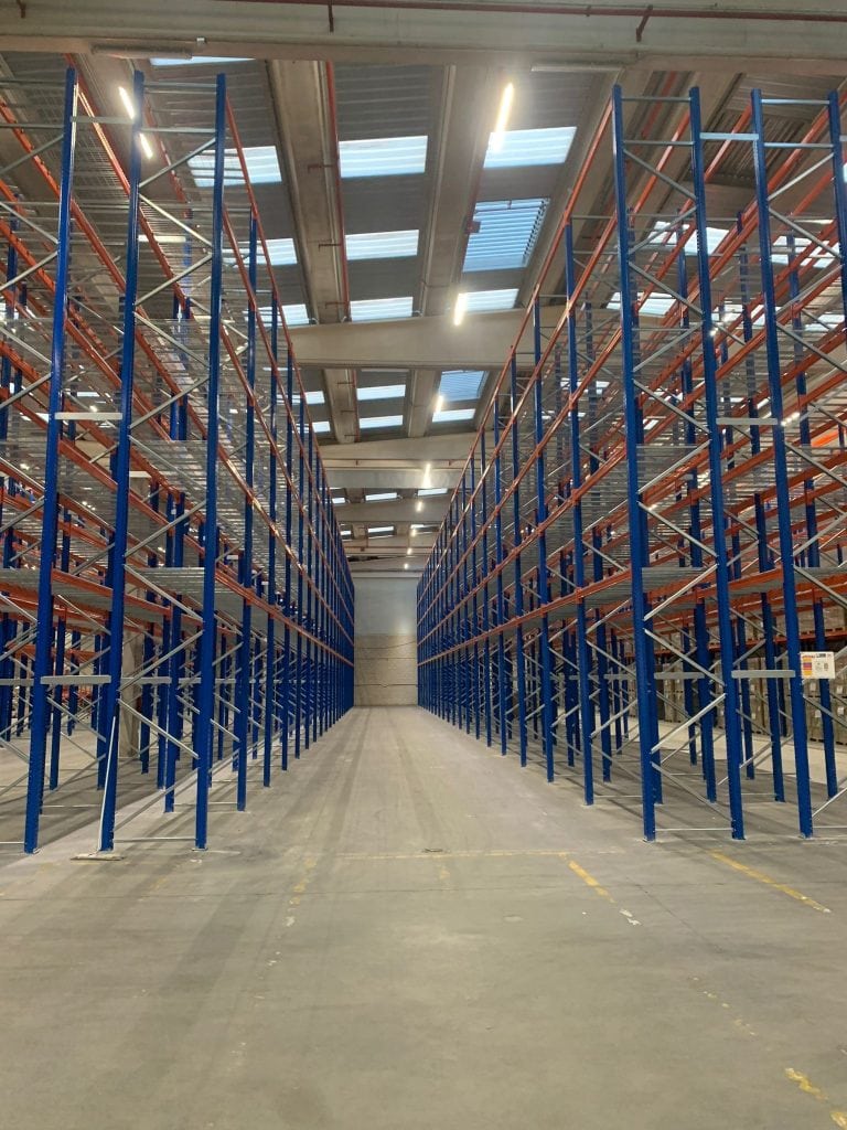 Empty warehouse with tall blue and orange metal shelving racks, concrete floor, and skylights in the ceiling.