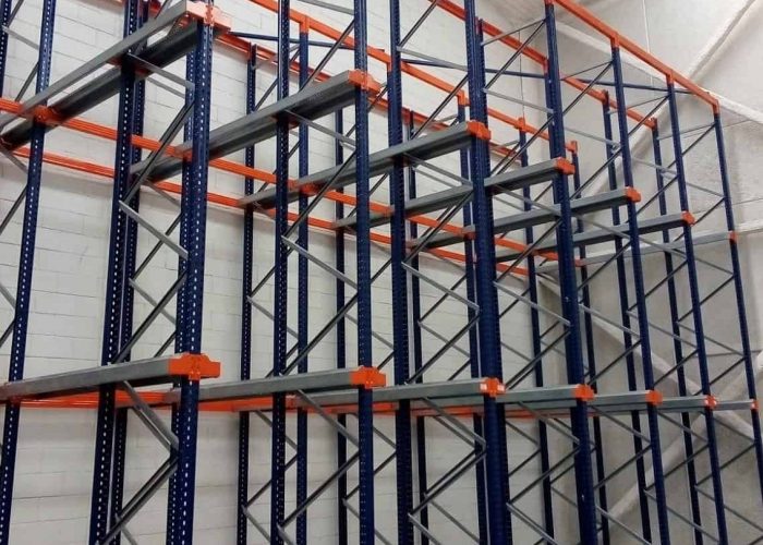Empty industrial storage racks with blue frames and orange supports are installed against a white brick wall in a warehouse with a high ceiling.