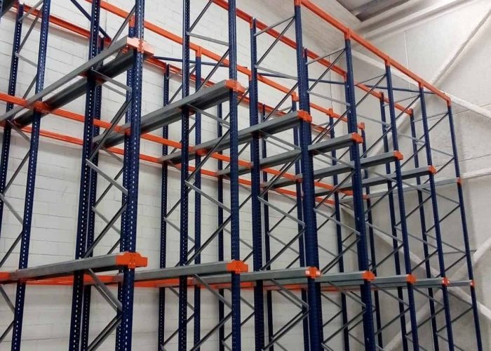 Empty industrial storage racks with blue frames and orange supports are installed against a white brick wall in a warehouse with a high ceiling.