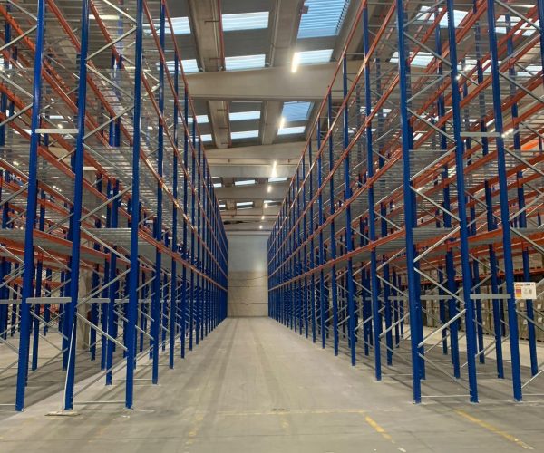 Empty warehouse with tall blue and orange metal shelving racks, concrete floor, and skylights in the ceiling.